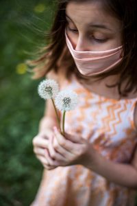 young woman, wearing a mask, holding fluffy white dandelions as if to blow them.